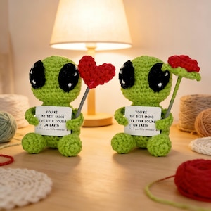 Peut inclure: Deux figurines d'extraterrestres verts crochetés avec de grands yeux noirs. L'une tient un cœur rouge, l'autre une fleur rouge. Chacune tient une pancarte avec l'inscription "YOU'RE THE BEST THING I'VE EVER FOUND ON EARTH".