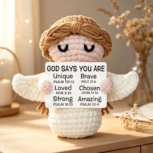 May include: A crocheted angel figurine with a light brown halo and hair, holding a sign that reads "GOD SAYS YOU ARE Unique, Brave, Loved, Chosen, Strong, Amazing." The angel has white wings and is made of soft yarn.