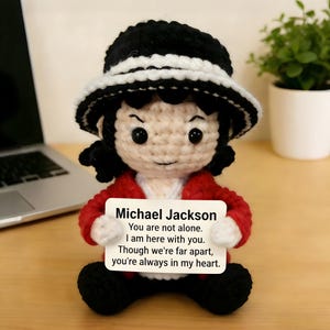 May include: Crocheted doll of Michael Jackson wearing a black hat with a white band, a red jacket, and black pants. The doll holds a sign with the text "Michael Jackson" and a message about being there for someone.
