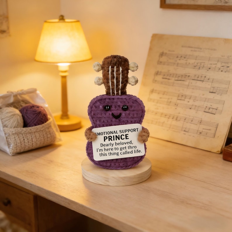 May include: A handmade purple crochet guitar with a smiling face and a sign that reads "EMOTIONAL SUPPORT PRINCE." The guitar is on a wooden base. The text on the sign says "Dearly beloved, I'm here to get thro this thing called life."