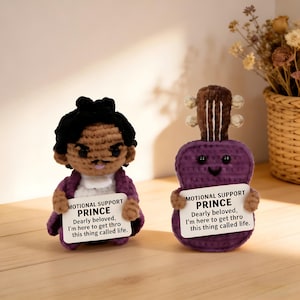 May include: Two handmade crochet dolls, one resembling a person and the other a purple guitar. Both dolls hold signs that read "MOTIONAL SUPPORT PRINCE" and a quote. The person doll has black hair and a purple jacket.