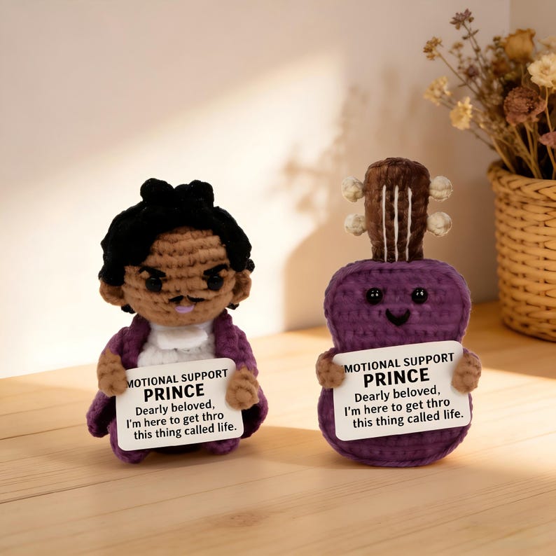 May include: Two handmade crochet dolls, one resembling a person and the other a purple guitar. Both dolls hold signs that read "MOTIONAL SUPPORT PRINCE" and a quote. The person doll has black hair and a purple jacket.