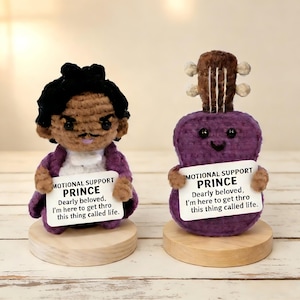 May include: Two handmade amigurumi figures: one resembling Prince, the other a purple guitar. Both hold signs that read "MOTIONAL SUPPORT PRINCE." The Prince figure wears a purple coat. The guitar has a smiling face.