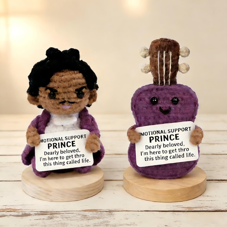 May include: Two handmade amigurumi figures: one resembling Prince, the other a purple guitar. Both hold signs that read "MOTIONAL SUPPORT PRINCE." The Prince figure wears a purple coat. The guitar has a smiling face.