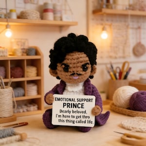 May include: A handmade crochet doll of Prince, wearing a purple jacket and holding a sign that reads "EMOTIONAL SUPPORT PRINCE." The doll has black hair and a mustache. The sign says "Dearly beloved, I'm here to get thro this thing called life."