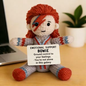 May include: A handmade crochet doll with red hair and a lightning bolt on its face, holding a sign that reads "EMOTIONAL SUPPORT BOWIE". The doll is wearing a gray outfit with red and blue accents. The text on the sign says "Ground control to your feelings. -You're not alone in this galaxy."