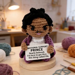 May include: A handmade crochet doll of Prince, wearing a purple jacket and white shirt, holding a sign that reads "MOTIONAL SUPPORT PRINCE." The doll has black hair and a mustache. It is displayed on a wooden base, with yarn and crochet hooks in the background.