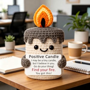 Puede incluir: Una vela de ganchillo con cuerpo color crema, cera gris que gotea en la parte superior y una llama naranja y amarilla. Tiene ojos de cuentas negras, una sonrisa y sostiene un cartel blanco que dice: Positive Candle, I may be a tiny candle, but I believe in you. Go do your thing! Find your fire. You got this!