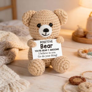May include: A crocheted teddy bear in tan yarn holds a sign that reads "POSITIVE Bear, You're Bear-y Awesome, I believe in you. Go do your thing!" The bear has black eyes and a white muzzle.