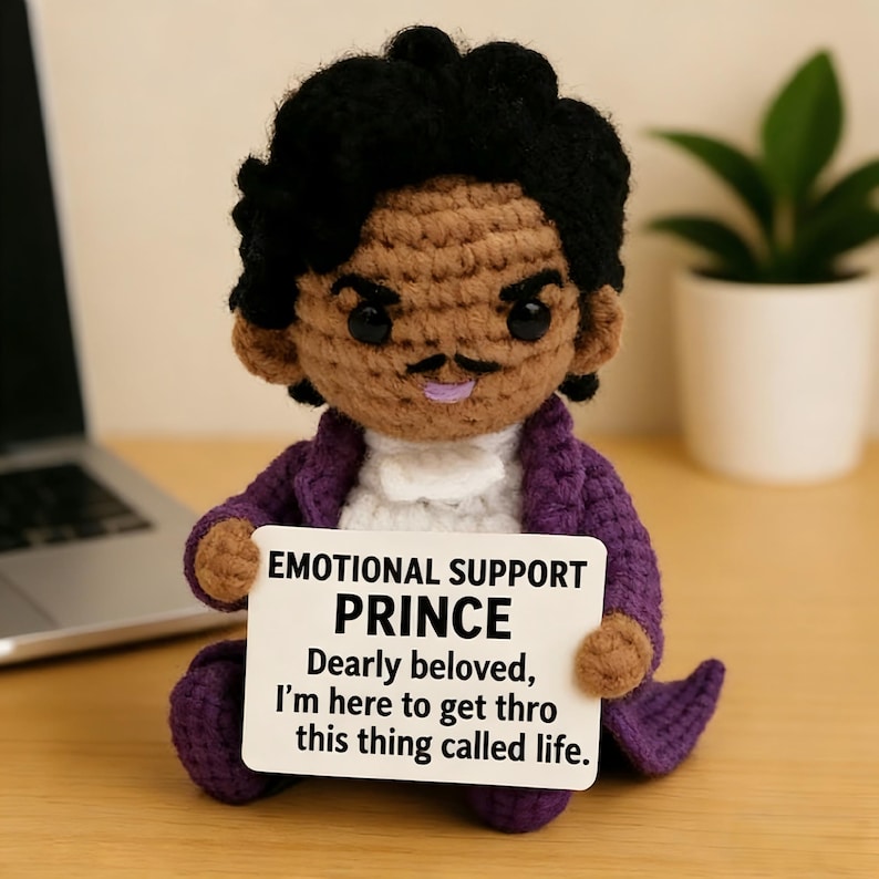 May include: A handmade crochet doll of Prince, wearing a purple jacket and holding a sign. The sign reads "EMOTIONAL SUPPORT PRINCE Dearly beloved, I'm here to get thro this thing called life."