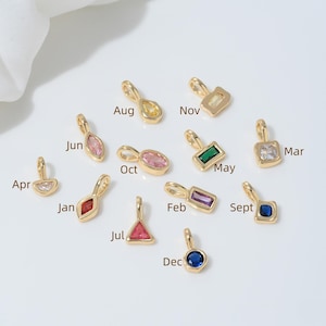 Peut inclure: Une collection de pendentifs dorés, chacun orné d'une pierre précieuse de forme différente et de couleurs variées. Chaque pendentif est doté d'une petite boucle en haut et est étiqueté avec un mois de l'année, comme "Août" et "Juin".