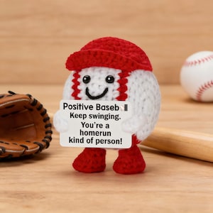 May include: A crocheted baseball character with a red cap and feet, holding a sign that reads "Positive Baseball. Keep swinging. You're a homerun kind of person!" A baseball glove and bat are in the background.