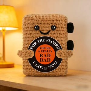Peut inclure: Un lecteur de disques en crochet beige fait à la main avec un visage souriant et des yeux en bouton noirs. Un autocollant circulaire sur le devant indique "FOR THE RECORD YOU'RE A REALLY RAD DAD I LOVE YOU."