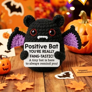 May include: A black crocheted bat plush toy with purple inner wings, large red eyes, and two white fangs. The bat holds a white sign that reads: Positive Bat YOU'RE REALLY FANG-TASTIC! A tiny bat is here to always remind you!