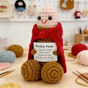 Könnte beinhalten: Gehäkelte Penis-Figur aus beige-braunem Garn, mit rotem Umhang und Schild. Auf dem Schild steht "Positive Penis" und der Text "I may be a tiny Penis, but I believe in you, Go do your thing!". Schwarze Knopfaugen und ein lächelnder Mund.