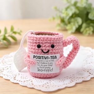 Puede incluir: Una taza de té rosa tejida a ganchillo con una cara sonriente y brazos, sosteniendo un letrero que dice "POSITIVI-TEA". El letrero incluye el texto "I may be a cup of tea, but I believe in you. Go do your thing! You're pretty Tea-riffic!"