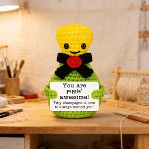 May include: A crocheted champagne bottle figure with a yellow head, green body, and black bow tie. It holds a sign that reads "You are poppin' awesome! Tiny champagne is here to always remind you!"