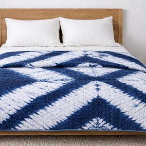 May include: A bed with a wooden frame and headboard, featuring a blue and white patterned quilt. Two white pillows rest on the bed. A small wooden side table with a lamp and a woven basket are also visible.