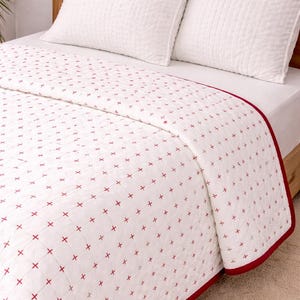 Op de afbeelding: Witte quilt met een rood kruissteekpatroon en een rode rand. Twee witte, getextureerde kussens liggen op een houten bedframe. Het beddengoed is netjes opgemaakt, wat een strakke uitstraling creëert. Deze quilt is ideaal voor een slaapkamer.