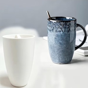 May include: A tall, cylindrical white ceramic cup with a smooth finish stands beside a blue and black speckled ceramic mug. The blue mug features a dark, glossy rim and a matching handle, with a small metal spoon resting inside the vessel.