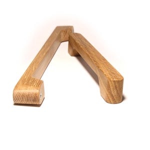 Può includere: Due maniglie per mobili in legno di rovere con finitura naturale. Le maniglie rettangolari hanno bordi arrotondati e fori per le viti. La venatura del legno è visibile. Le maniglie sono su sfondo bianco. Ogni maniglia è lunga circa 15 cm.