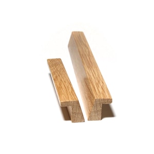 May include: Two wooden cabinet pulls with a natural oak finish. The pulls have a T-shaped design and are set against a white background. The wood grain is visible, adding texture to the minimalist design.