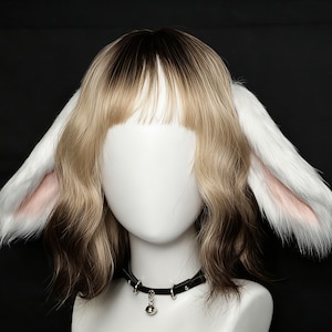 Puede incluir: Una peluca a la altura de los hombros con raíces marrón oscuro que pasan a puntas rubio claro, con flequillo recto y largas orejas de animal de peluche blancas con detalles rosados. Se lleva una gargantilla negra con un pequeño cascabel plateado.
