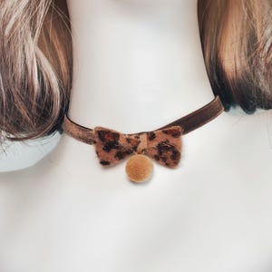 May include: A brown velvet choker necklace featuring a central bow with a brown and black leopard print pattern. A small, round, tan-colored fuzzy pom-pom charm hangs from the center of the bow, resting against the neck.