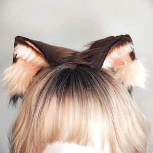 May include: A pair of faux fur animal ears in shades of dark brown, tan, and cream. The ears feature a pointed shape with dense, textured fur, lighter cream-colored tufts at the base, and a soft, multi-toned appearance with dark brown tips.