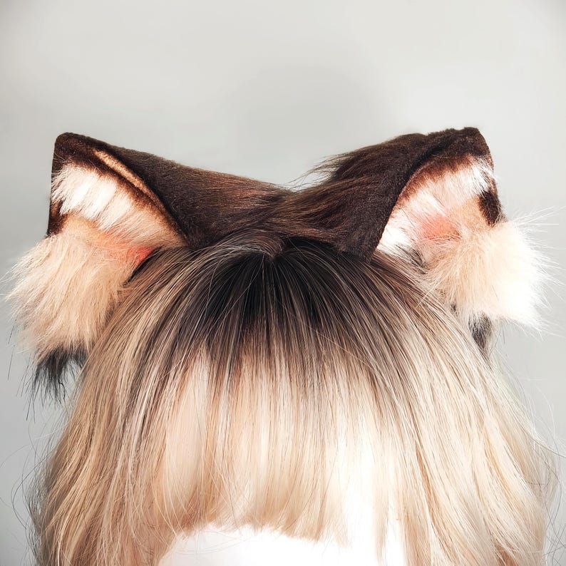 May include: A pair of faux fur animal ears in shades of dark brown, tan, and cream. The ears feature a pointed shape with dense, textured fur, lighter cream-colored tufts at the base, and a soft, multi-toned appearance with dark brown tips.