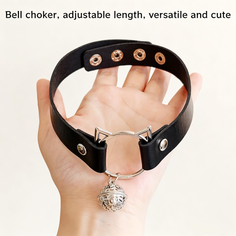 K&ouml;nnte beinhalten: Ein schwarzes Halsband mit einem silberfarbenen Metallring, Katzenohr-Akzenten und einem h&auml;ngenden Filigran-Glockenanh&auml;nger. Das Band verf&uuml;gt &uuml;ber vier verstellbare Druckkn&ouml;pfe. Der Text "Bell choker, adjustable length, versatile and cute" ist sichtbar.