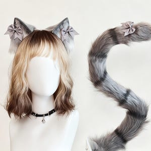 May include: A set of faux fur cat ears on a headband and a matching long striped tail. The ears are grey with white tips and small grey bows. The tail is grey and black with a small grey bow at the top. A black choker with a silver bell is included.