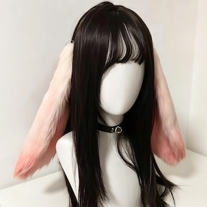 Könnte beinhalten: Eine lange, glatte, dunkelbraune Perücke mit Pony, die an den Seiten zwei große, flauschige Kunstfell-Hasenohren aufweist. Die Ohren verlaufen von einer hellen Cremefarbe an der Basis zu einem sanften Rosa an den Spitzen. Ein schwarzes Halsband mit einem silbernen Ring wird getragen.