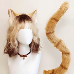 Op de afbeelding: Een pruik op schouderlengte met een blonde en bruine ombre-kleuring, voorzien van aangehechte kattenoren van imitatiebont. Een lange, bijpassende oranje en bruin gestreepte staart van imitatiebont staat ernaast. Een zwarte choker met strik is inbegrepen.