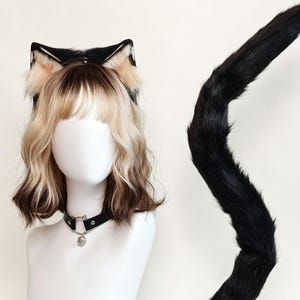 K&ouml;nnte beinhalten: Ein Set aus schwarzen und beigen Kunstfell-Katzenohren auf einem Haarreif, einem langen schwarzen Kunstfellschwanz und einem schwarzen Kunstleder-Halsband mit einem silberfarbenen Metallring und einem kleinen runden Anh&auml;nger.
