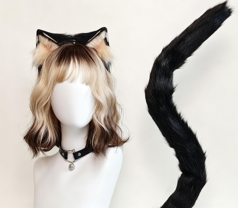 K&ouml;nnte beinhalten: Ein Set aus schwarzen und beigen Kunstfell-Katzenohren auf einem Haarreif, einem langen schwarzen Kunstfellschwanz und einem schwarzen Kunstleder-Halsband mit einem silberfarbenen Metallring und einem kleinen runden Anh&auml;nger.