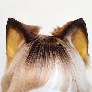 Può includere: Un paio di orecchie da animale in pelliccia sintetica nei toni del marrone, beige e nero, caratterizzate da una forma a punta con bordi scuri e un interno marrone dorato, fissate su una base di capelli lisci e scalati con riflessi biondi.
