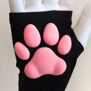 K&ouml;nnte beinhalten: Ein schwarzer fingerloser Handschuh mit einem erhabenen, weichen, rosafarbenen Silikon-Pfotenabdruck auf der R&uuml;ckseite. Der Handschuh besteht aus einem glatten, dehnbaren Stoff und zeigt vier kleine ovale Zehenballen &uuml;ber einem gro&szlig;en, herzf&ouml;rmigen Ballen.