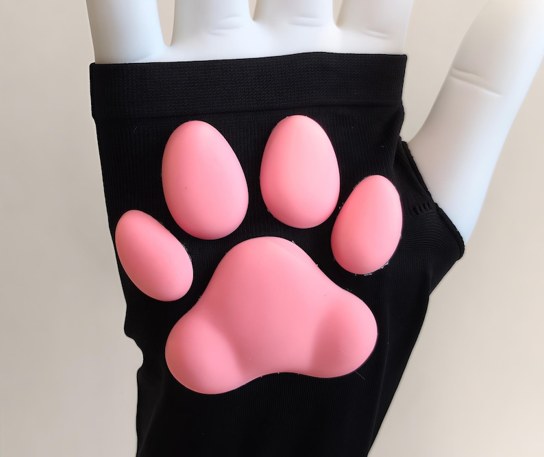 K&ouml;nnte beinhalten: Ein schwarzer fingerloser Handschuh mit einem erhabenen, weichen, rosafarbenen Silikon-Pfotenabdruck auf der R&uuml;ckseite. Der Handschuh besteht aus einem glatten, dehnbaren Stoff und zeigt vier kleine ovale Zehenballen &uuml;ber einem gro&szlig;en, herzf&ouml;rmigen Ballen.