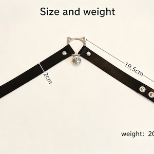 K&ouml;nnte beinhalten: Ein schwarzes Halsband aus leder&auml;hnlichem Material mit silberfarbenen Metallbeschl&auml;gen, einem katzenohrf&ouml;rmigen Ring und einer kleinen runden Glocke. Das Band ist 2cm breit und 19,5cm lang, bei einem Gewicht von 20g. Text Size and weight sichtbar.