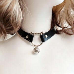 K&ouml;nnte beinhalten: Ein schwarzes Lederhalsband mit einem silberfarbenen Metallring, der oben zwei kleine spitze Katzenohren aufweist. Eine runde, filigran gemusterte silberne Glocke h&auml;ngt in der Mitte des Rings. Das Halsband ist mit zwei silbernen Nieten befestigt.