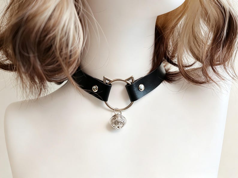 K&ouml;nnte beinhalten: Ein schwarzes Lederhalsband mit einem silberfarbenen Metallring, der oben zwei kleine spitze Katzenohren aufweist. Eine runde, filigran gemusterte silberne Glocke h&auml;ngt in der Mitte des Rings. Das Halsband ist mit zwei silbernen Nieten befestigt.