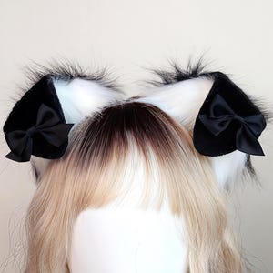 May include: Black and white cat ears with a fluffy texture and black satin bows. The ears are attached to a headband and are designed to be worn on the head. The ears are a cosplay accessory.