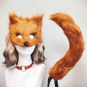 May include: A fox mask made of orange faux fur with a black nose, paired with a matching long orange faux fur tail. The set includes a brown leather-like choker collar featuring a central cat-shaped metal ring and two small silver bells.