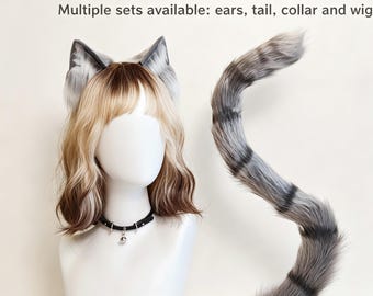 May include: A set featuring a blonde and brown wig on a mannequin head, topped with grey and white faux fur cat ears. A black collar with a small silver bell is around the neck. A long, grey and white striped faux fur tail is positioned to the side.