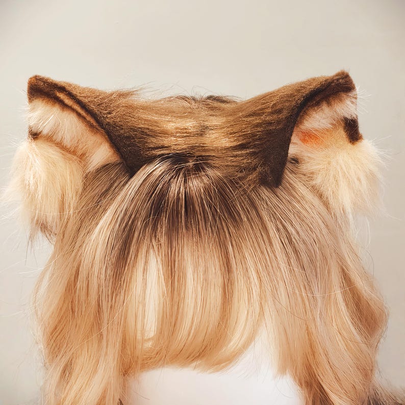 May include: A pair of faux fur animal ears in shades of brown, tan, and cream. The ears feature dark brown pointed tips, a textured brown center, and long, fluffy cream-colored fur accents at the base, attached to a light brown wig with bangs.
