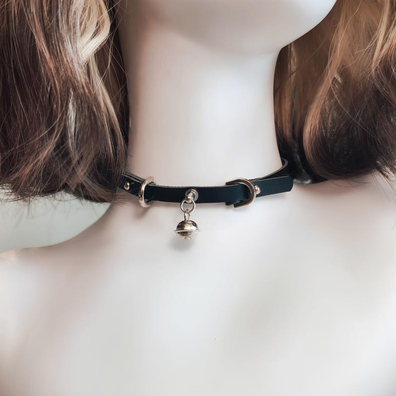K&ouml;nnte beinhalten: Ein schwarzes Lederhalsband mit einer silberfarbenen Metallschnalle, einem kreisf&ouml;rmigen Ring und einem kleinen, h&auml;ngenden Glockenanh&auml;nger. Das Band wird mit einer Metalldornschnalle verschlossen und verf&uuml;gt &uuml;ber silberne Nieten am Riemen.