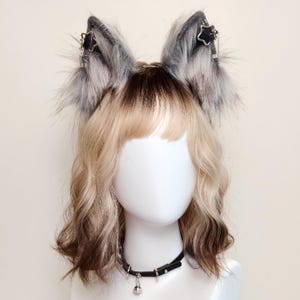 May include: A pair of gray and white faux fur cat ears with black star accents, attached to a headband. A black choker necklace with a silver bell and studs is also visible. The ears and necklace are displayed on a mannequin head with blonde and brown wavy hair.