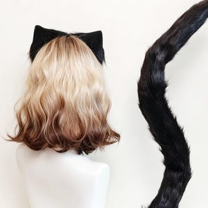 K&ouml;nnte beinhalten: Eine schulterlange Per&uuml;cke mit blondem Ansatz und braunen Spitzen, versehen mit schwarzen Katzenohren aus Kunstfell. Ein langer, geschwungener schwarzer Kunstfellschwanz ist rechts neben der Per&uuml;cke auf einer wei&szlig;en B&uuml;ste platziert.