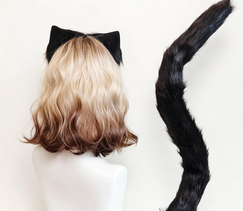 K&ouml;nnte beinhalten: Eine schulterlange Per&uuml;cke mit blondem Ansatz und braunen Spitzen, versehen mit schwarzen Katzenohren aus Kunstfell. Ein langer, geschwungener schwarzer Kunstfellschwanz ist rechts neben der Per&uuml;cke auf einer wei&szlig;en B&uuml;ste platziert.